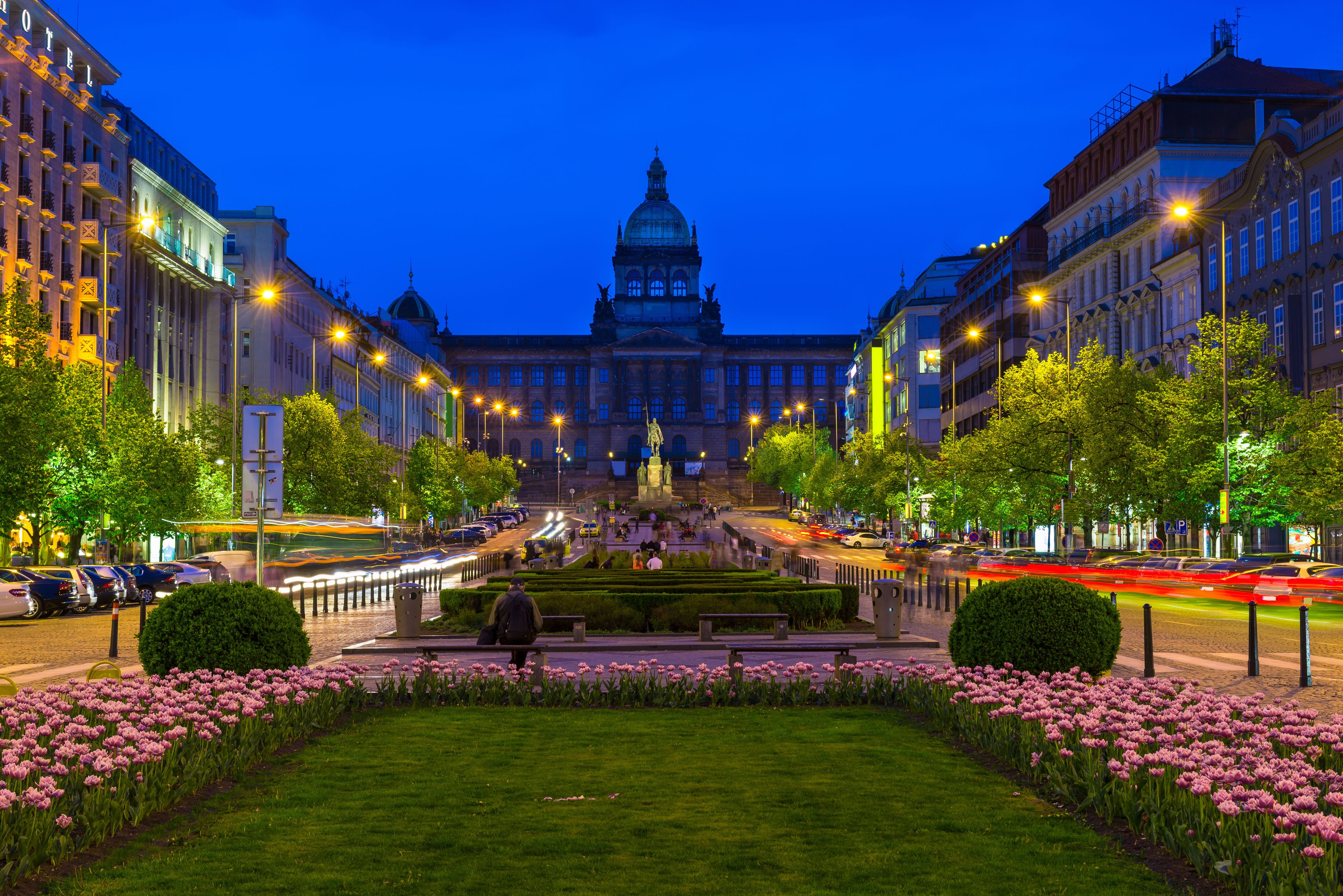 A city square with a large building in the background, surrounded by trees and flower beds.