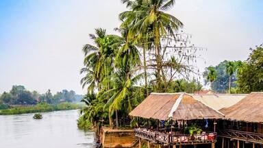 A calm river with a traditional boat docked on the right, surrounded by thatched-roof huts and palm trees.