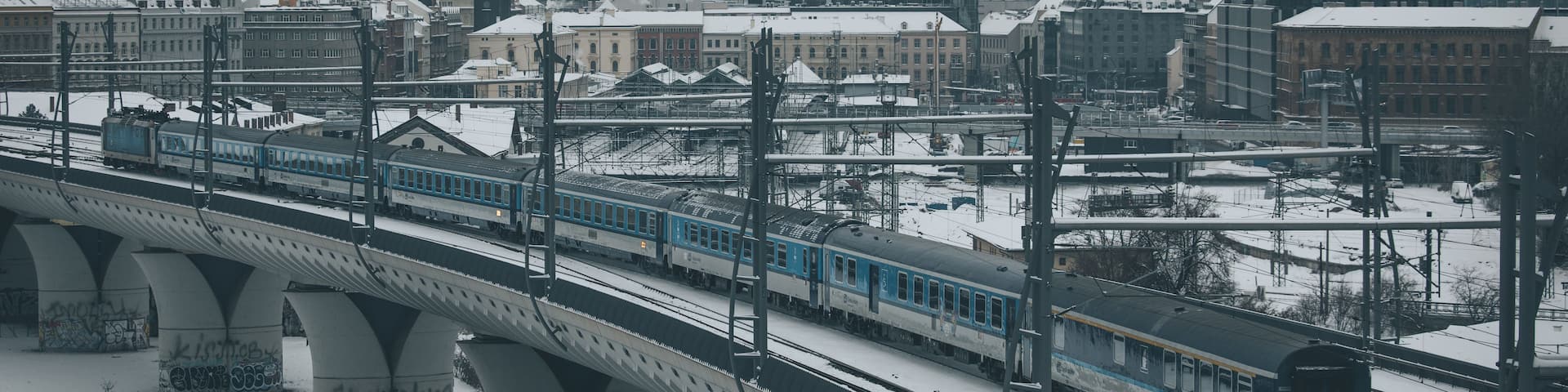 A train on a bridge over a river, with snow-covered buildings and a distant cityscape.