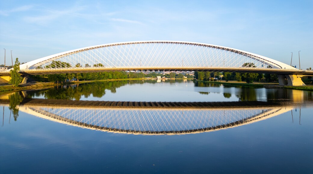 Troja Bridge stands over the Vltava River in Prague during sunrise. The calm waters reflect the structure, creating a clear view of the bridge and surroundings.