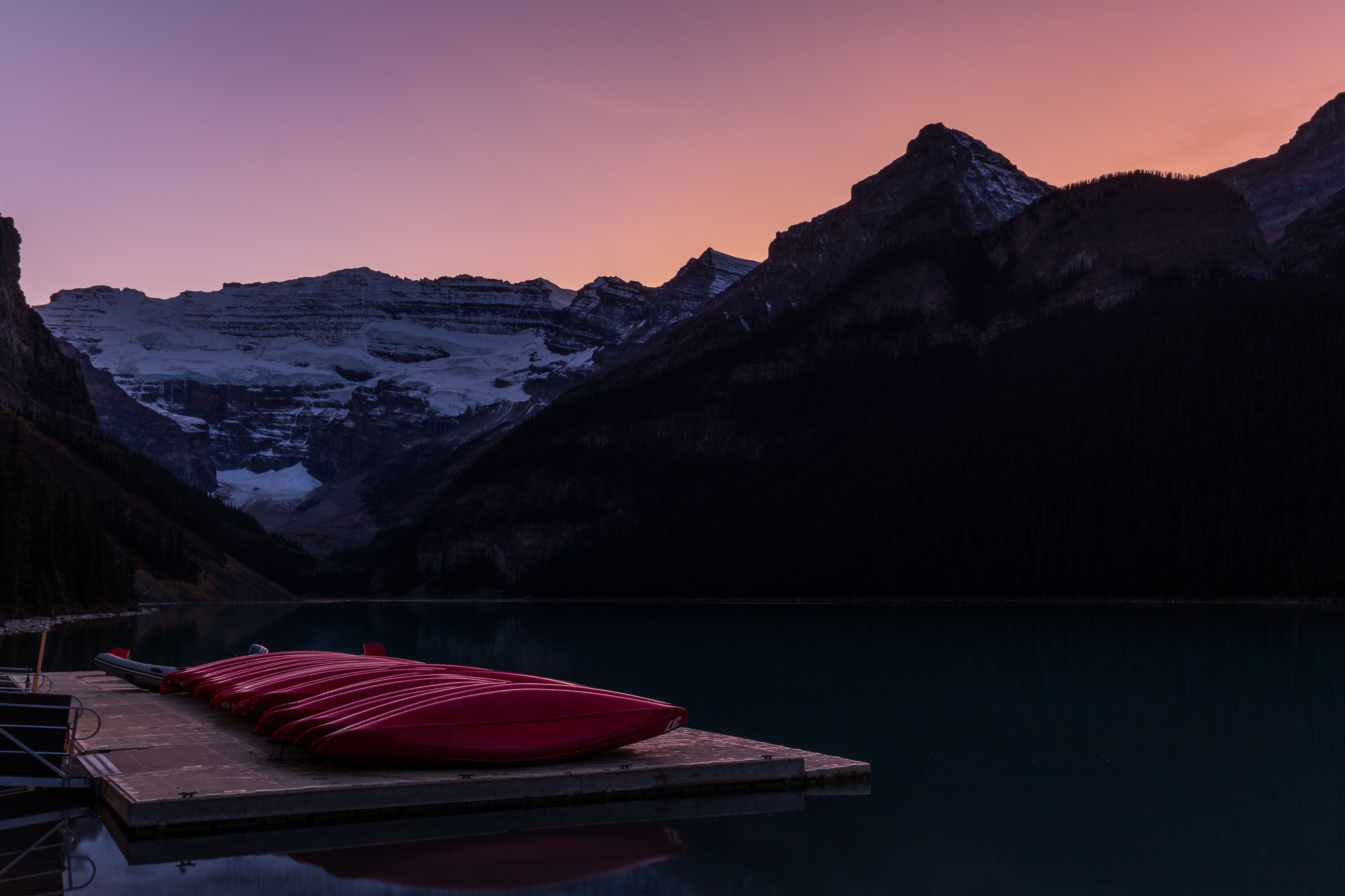 A wooden dock with red kayaks at a lakeside with mountains in the background.