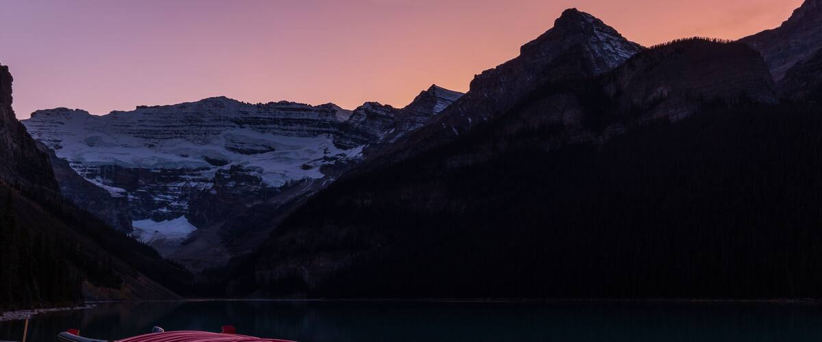 A wooden dock with red kayaks at a lakeside with mountains in the background.