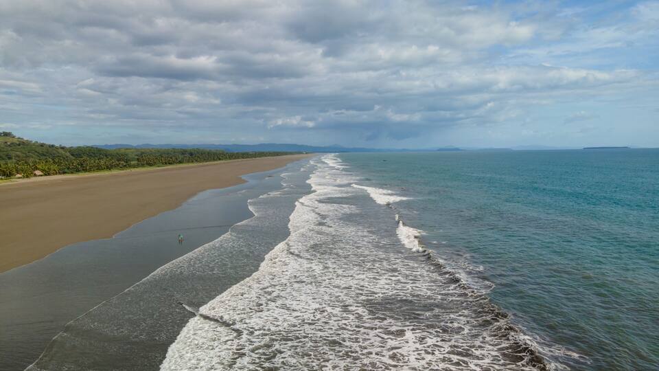 A beach with waves lapping onto the shore, a clear sky, and distant mountains.