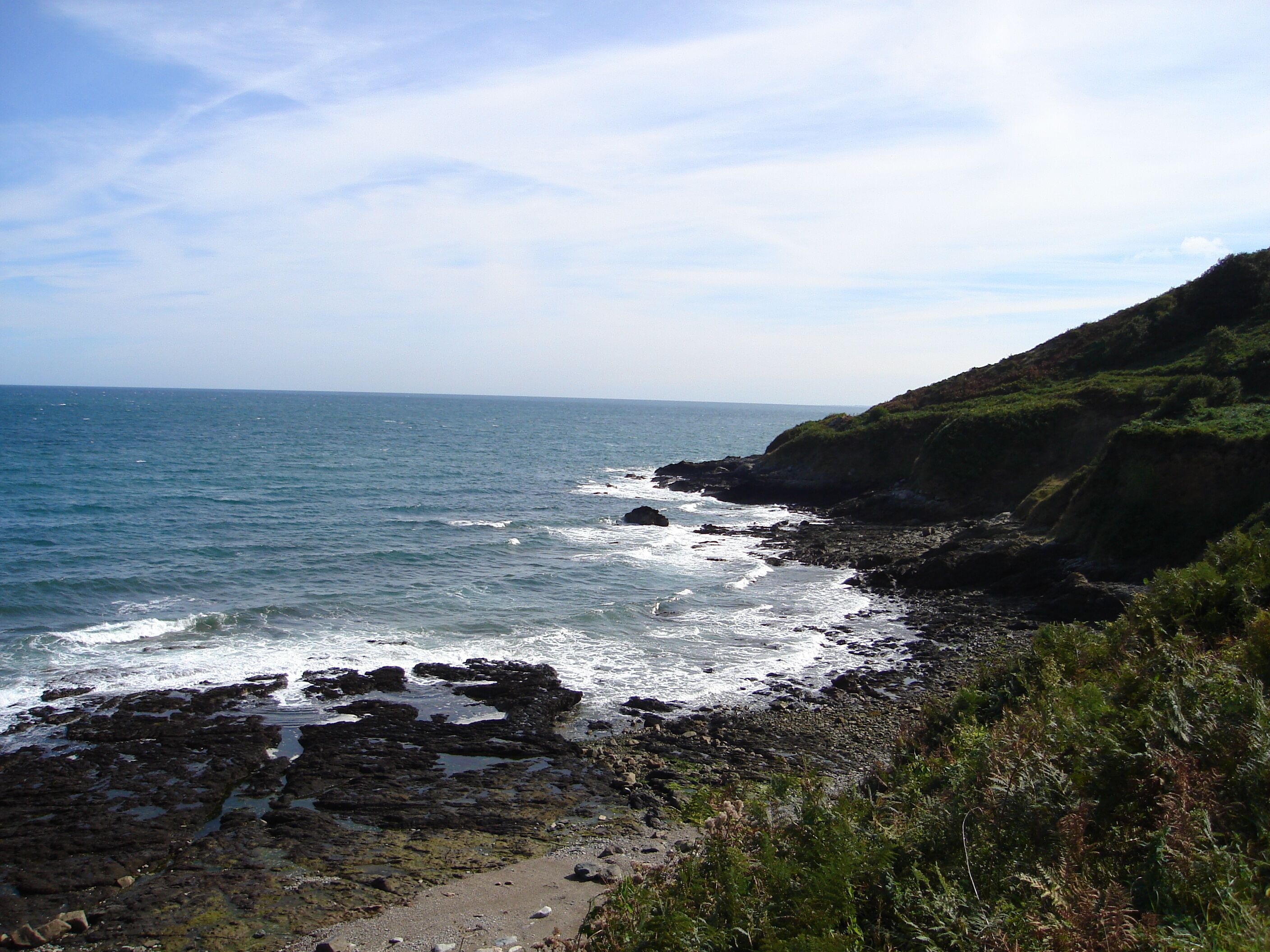 A rocky coastline with waves crashing against the shore, a clear sky, and a distant view of the ocean.