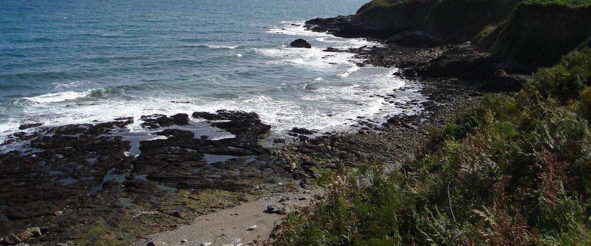 A rocky coastline with waves crashing against the shore, a clear sky, and a distant view of the ocean.