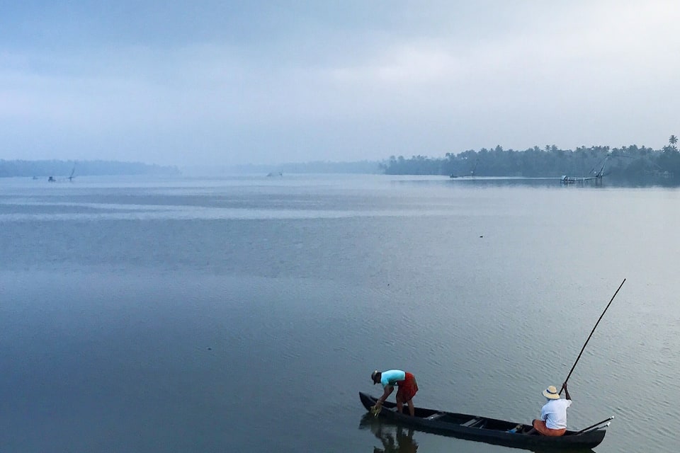 A calm lake with two people in a small boat, one standing and one sitting, fishing.