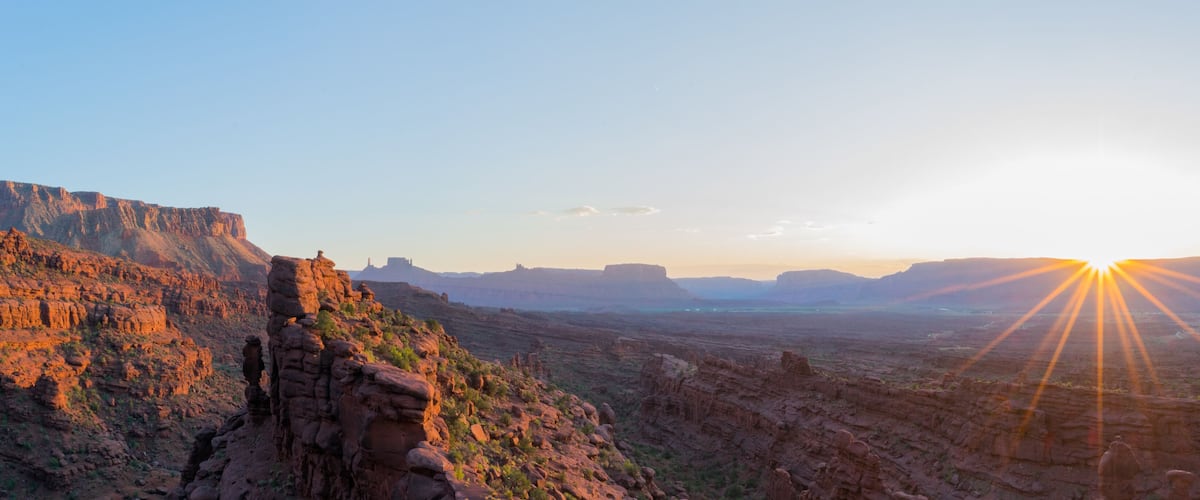 A canyon with a prominent rock formation on the left and the sun setting in the distance.