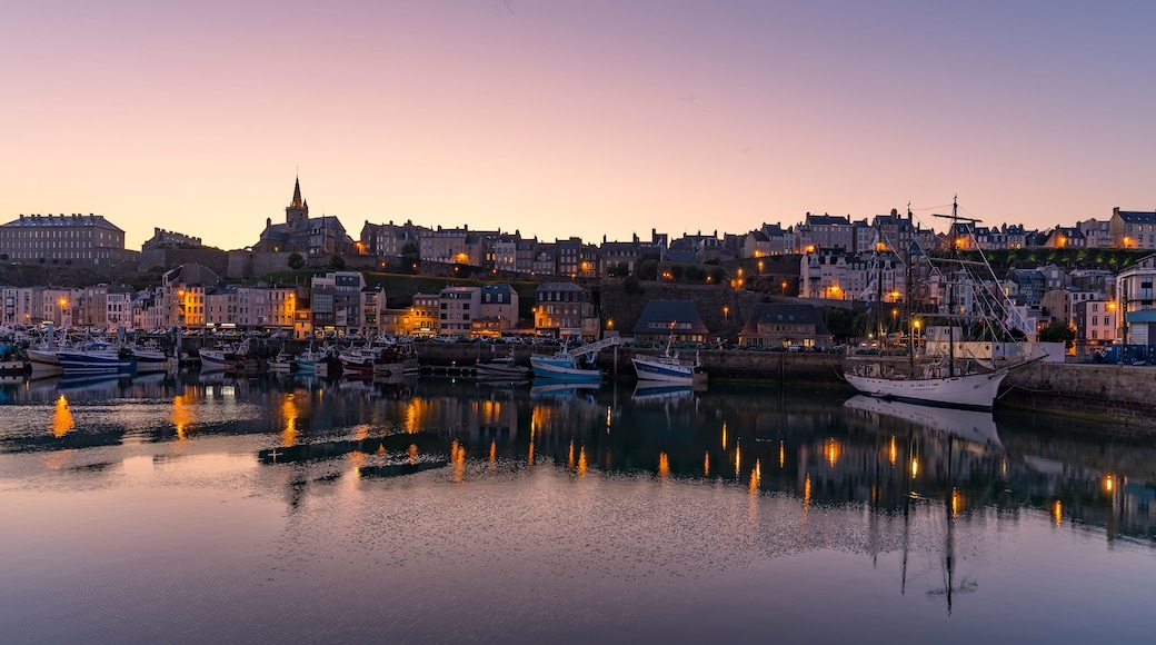 Une ville côtière au crépuscule, avec un port rempli de bateaux, des bâtiments historiques et le clocher imposant d’une église.