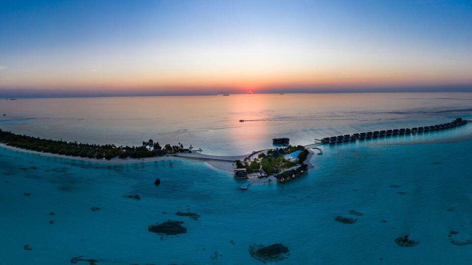 A tropical island at sunset with clear turquoise waters, a long pier, and a small cluster of buildings.