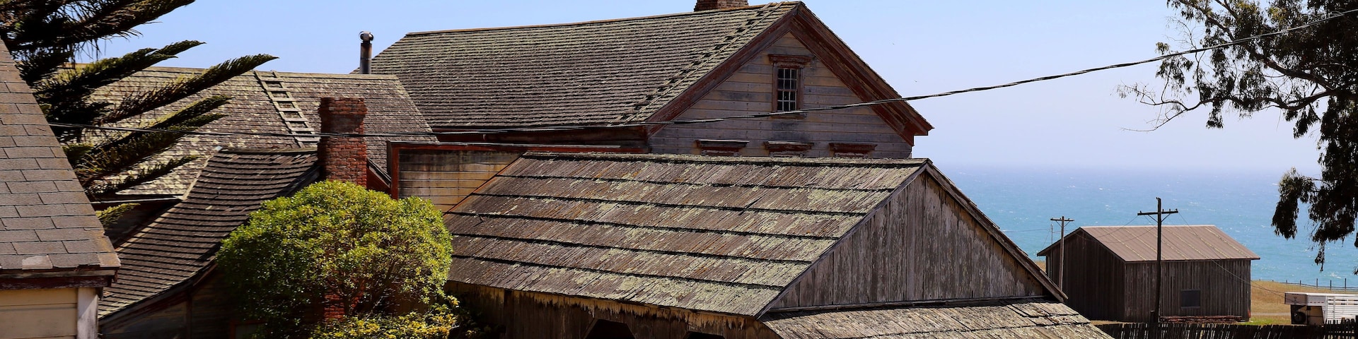 A rustic wooden building with a steeply pitched roof, surrounded by a fence and a variety of vegetation.