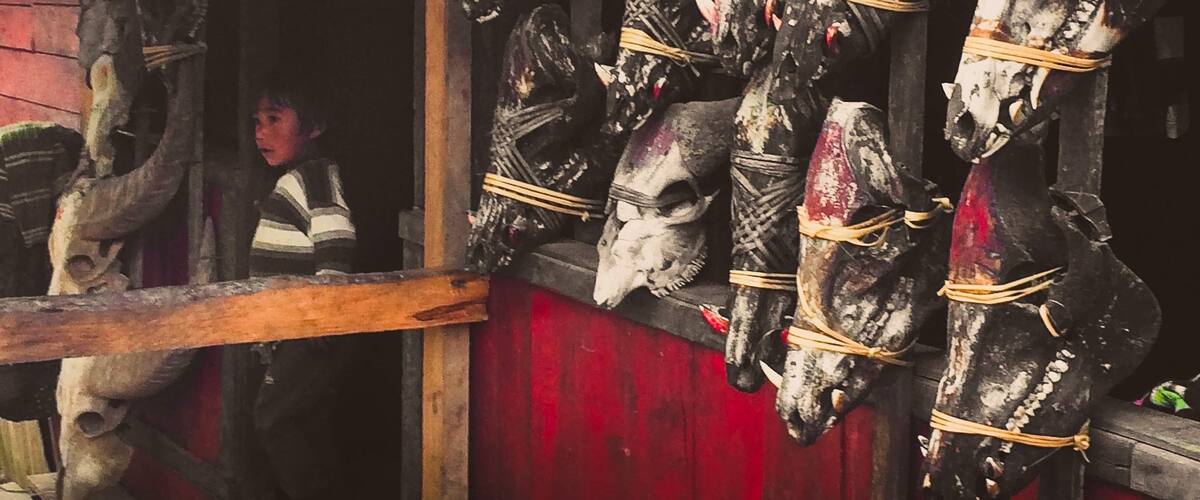 A collection of carved wooden figures, some with skull-like features, tied with strings, displayed against a red background.