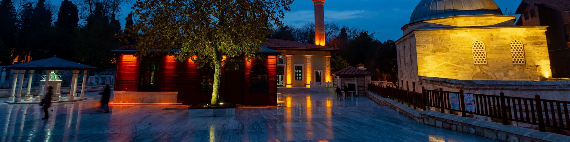 A mosque with a minaret and a dome, surrounded by a reflective marble courtyard and a large tree.