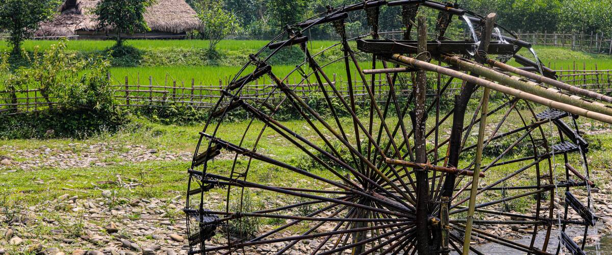 A traditional water wheel in a rural setting with lush greenery and a thatched-roof house in the background.