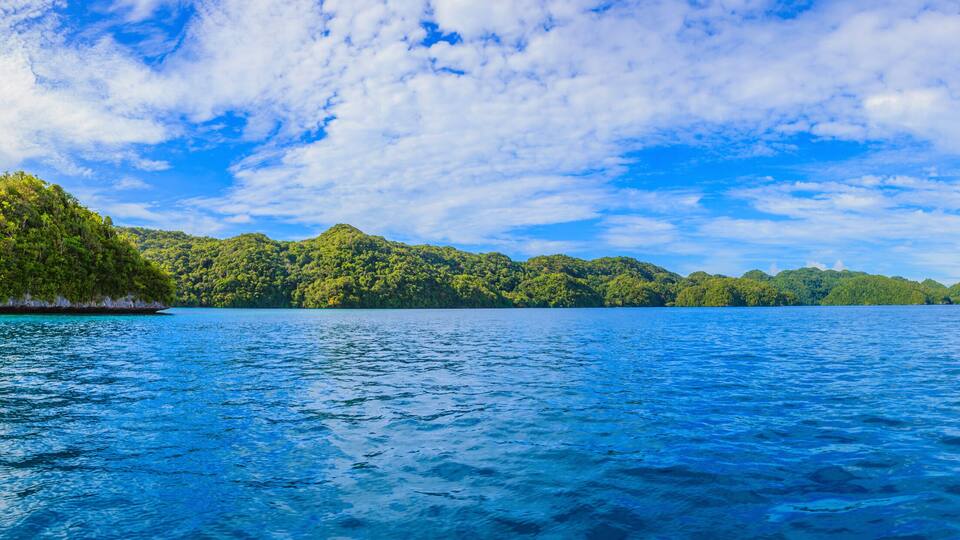 A tropical island surrounded by clear blue waters and lush green hills under a partly cloudy sky.