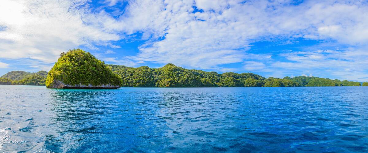 A tropical island surrounded by clear blue waters and lush green hills under a partly cloudy sky.