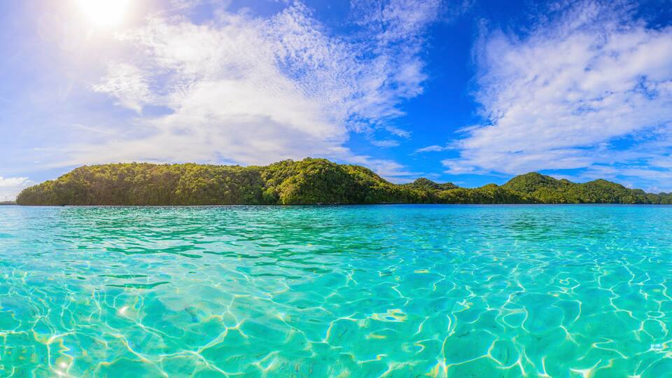 A clear turquoise sea with visible underwater patterns, surrounded by lush green islands and a bright blue sky with scattered clouds.