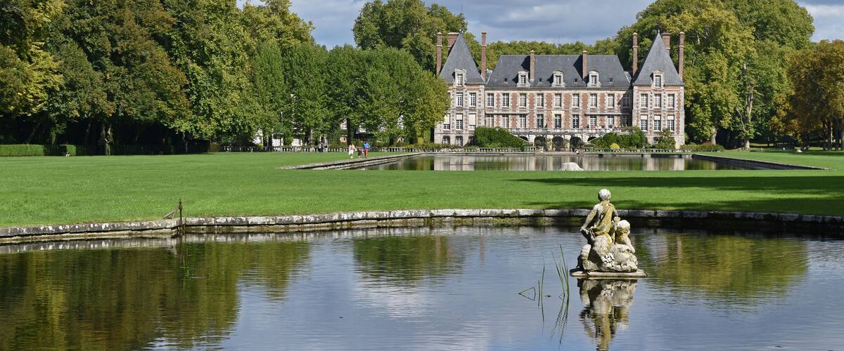 A large, historic building with a symmetrical facade and steeply pitched roofs, surrounded by lush green lawns and a calm pond with a statue in the foreground.