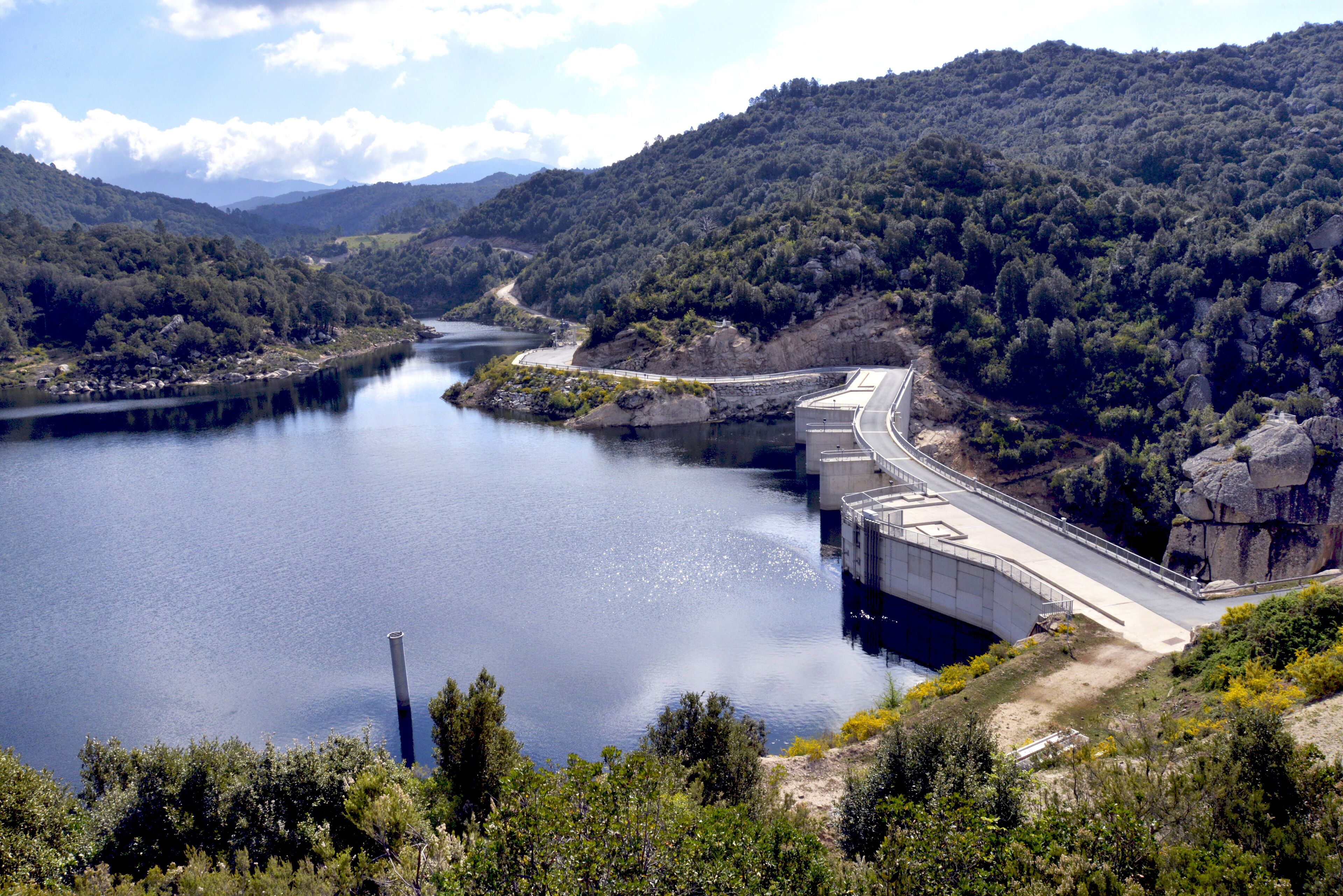 A dam with a road passing over it, surrounded by a forested valley and a body of water.