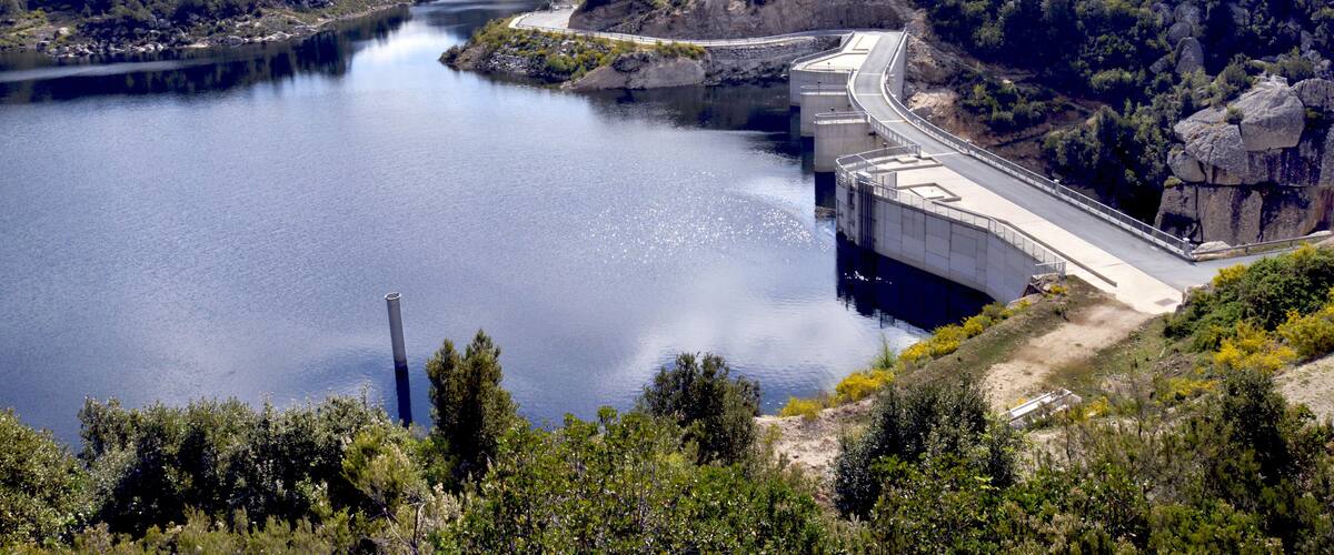 A dam with a road passing over it, surrounded by a forested valley and a body of water.