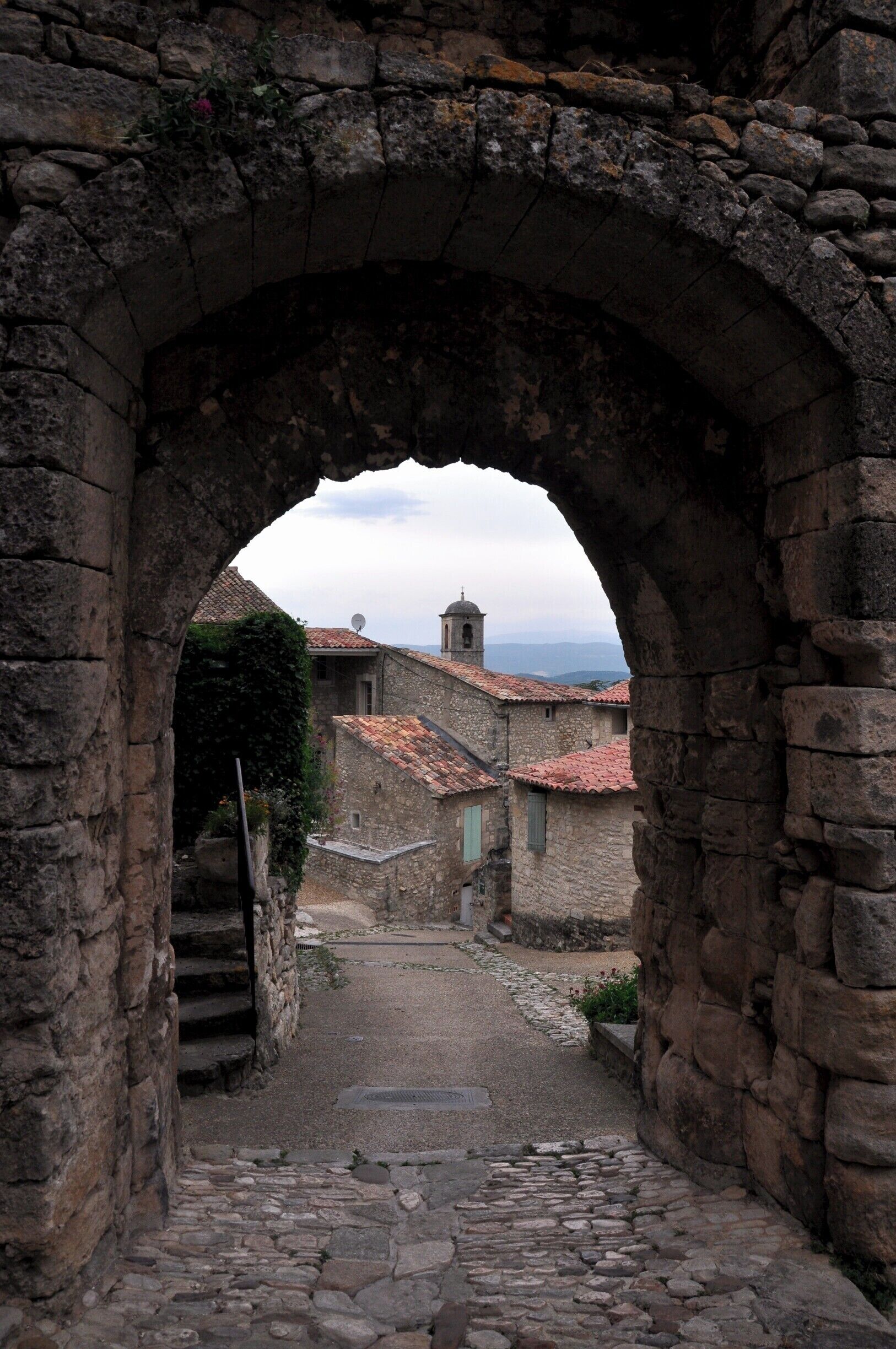 A stone archway leading to a narrow cobblestone path flanked by stone buildings with red-tiled roofs.