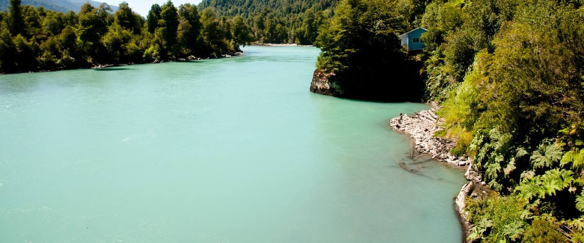 A turquoise river winding through a forested valley with a small house on a cliff.