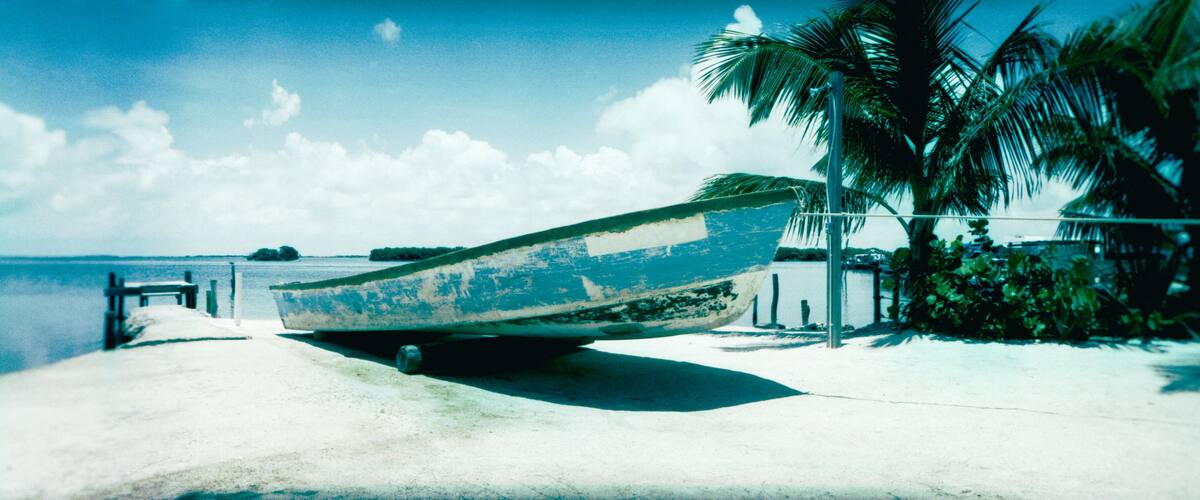 A weathered boat on a sandy beach with palm trees and a clear sky.
