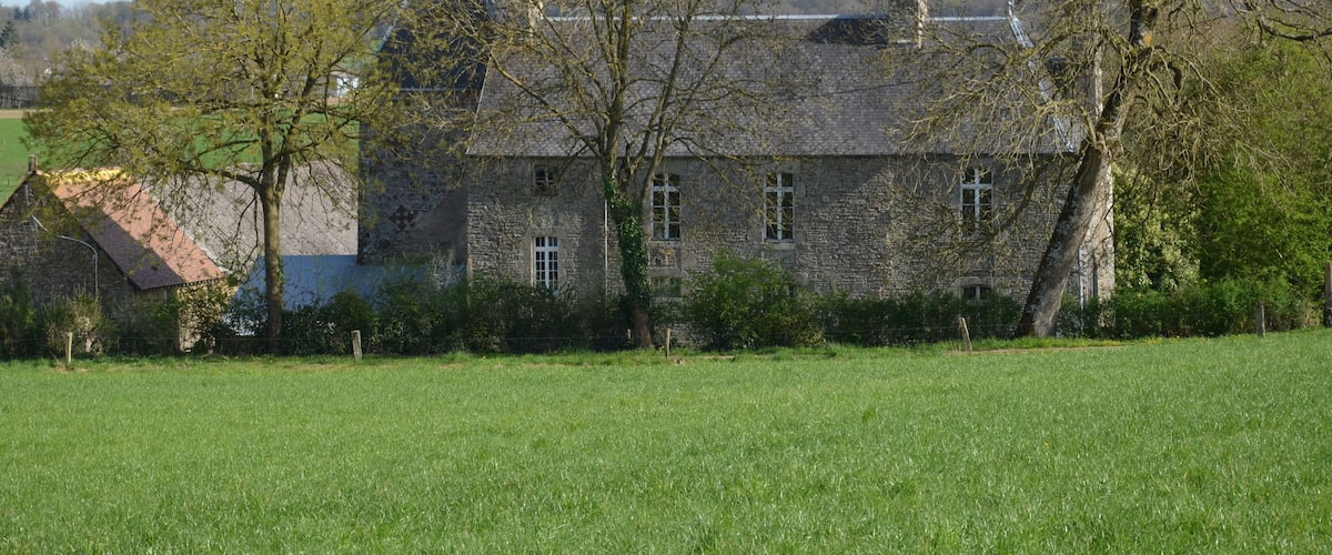 A stone house with a sloped roof, surrounded by a green field and trees.