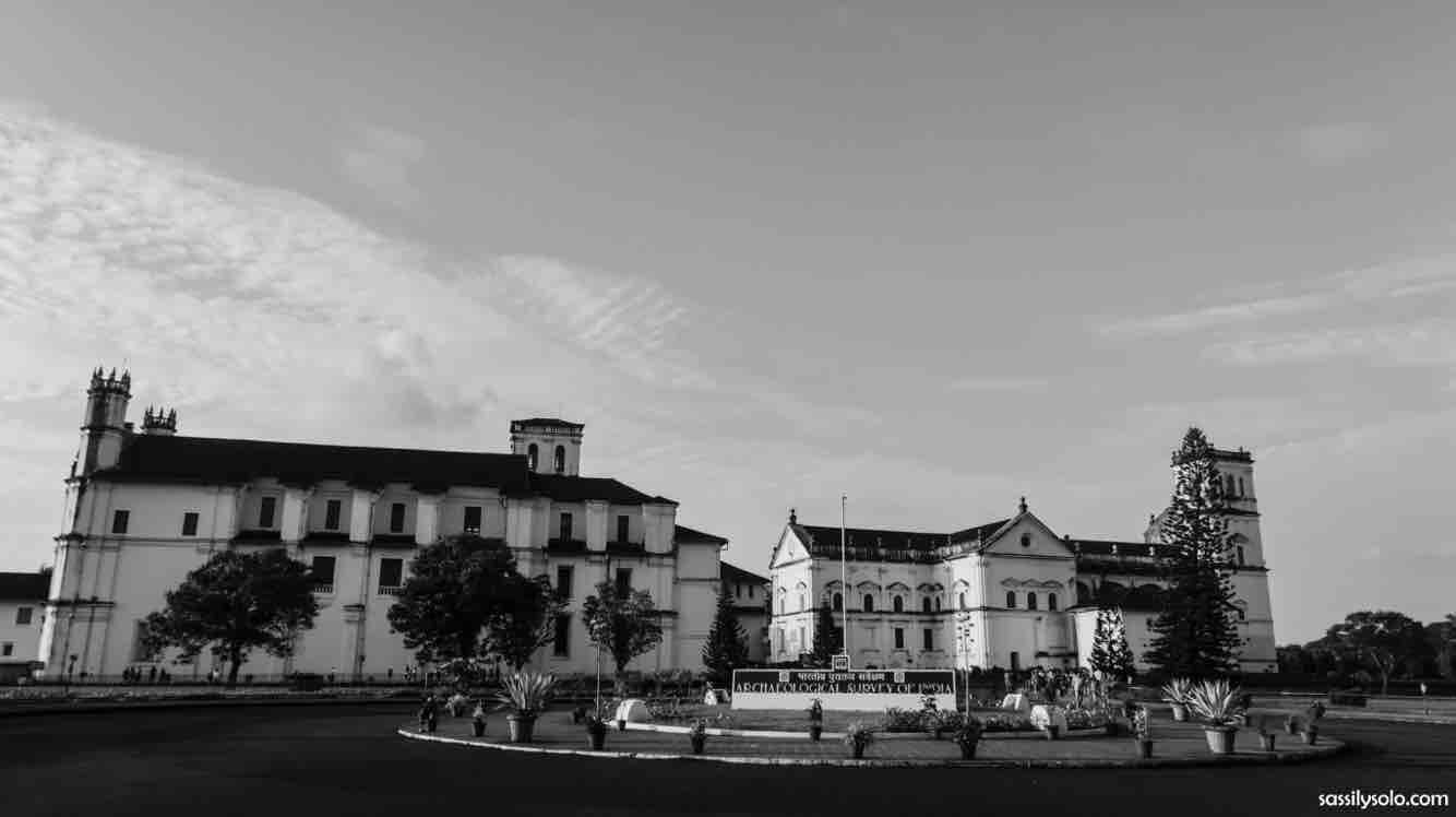 Um edifício histórico com uma torre do relógio imponente e uma grande torre central, cercado por um jardim paisagístico com árvores.