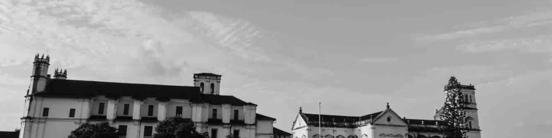 Um edifício histórico com uma torre do relógio imponente e uma grande torre central, cercado por um jardim paisagístico com árvores.
