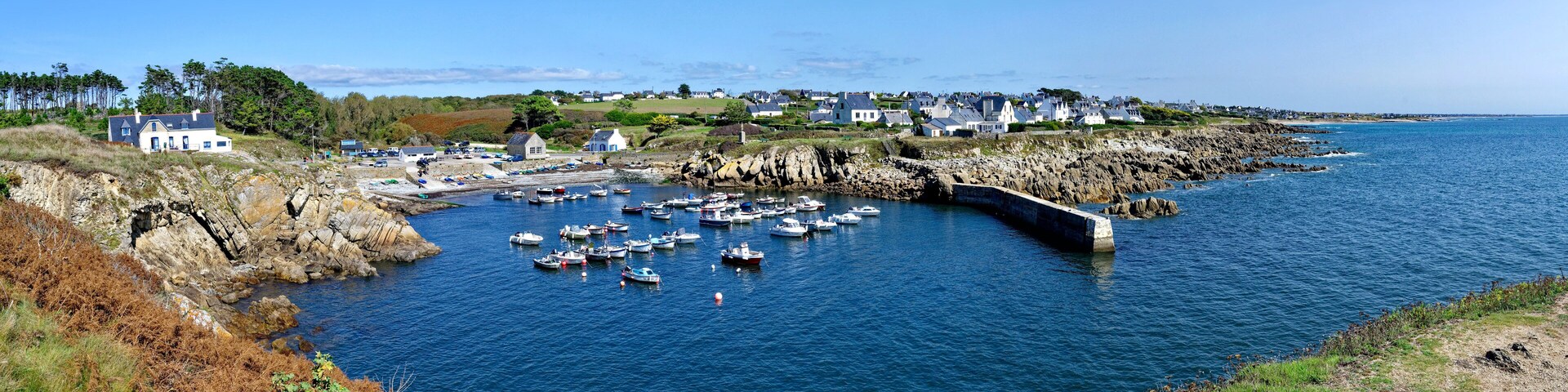 A coastal town with houses, a rocky shoreline, and numerous boats docked in a harbor.