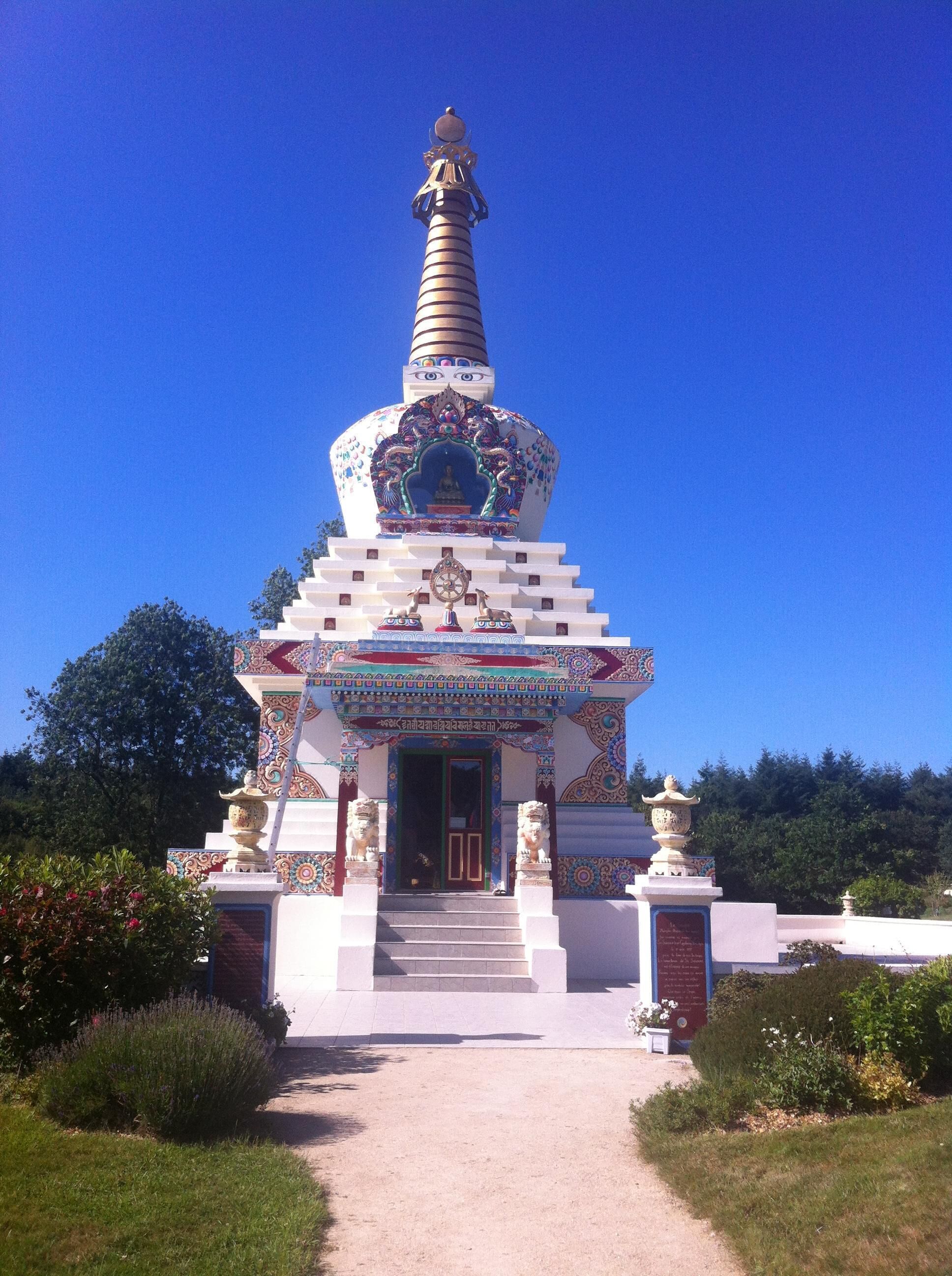 A colorful stupa with intricate designs and a statue on top, surrounded by a pathway and greenery.