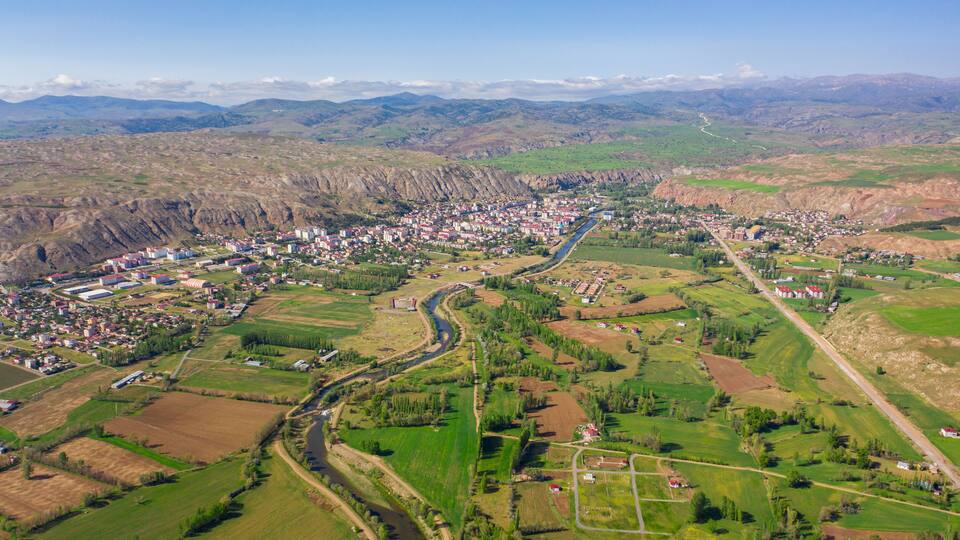 An aerial view of a town nestled in a valley with a river running through it, surrounded by green fields and mountains in the background.