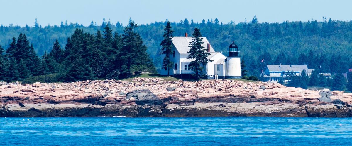 A coastal town with a lighthouse, rocky shoreline, dense forest, and a clear blue sky.