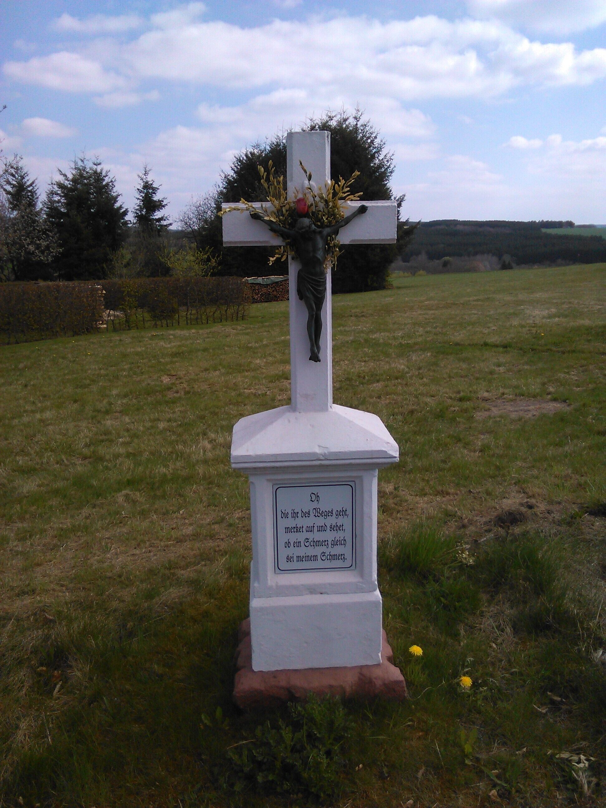 A white cross with a crucifix and flowers, surrounded by grass and trees.