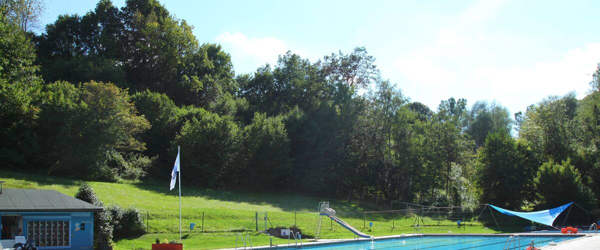 A swimming pool with a blue tarp shade, a small building, and a grassy area with trees in the background.