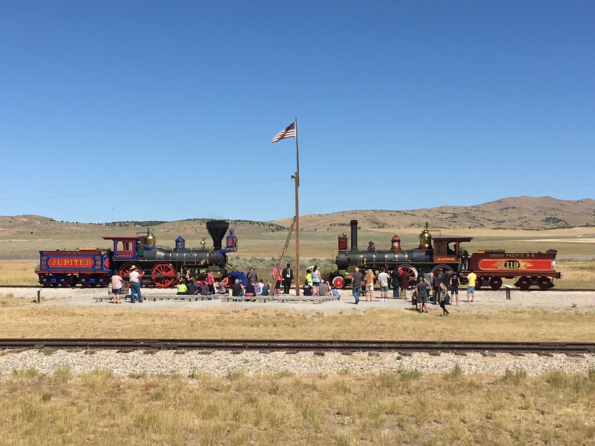 Two vintage steam locomotives, a Union Pacific RR 119 and a Jupiter, parked on a railway track with a crowd of people around them in a dry, hilly landscape.