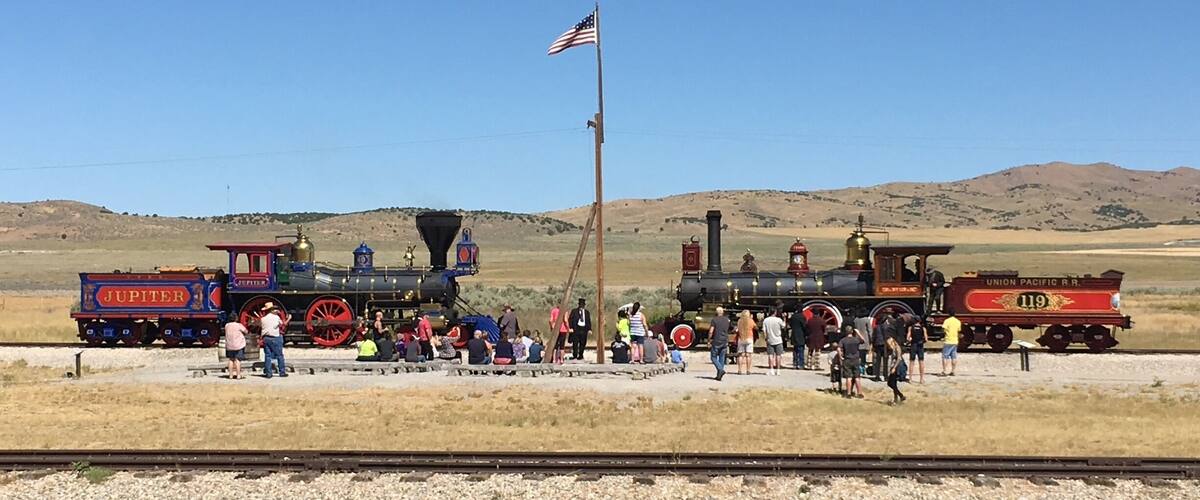 Two vintage steam locomotives, a Union Pacific RR 119 and a Jupiter, parked on a railway track with a crowd of people around them in a dry, hilly landscape.
