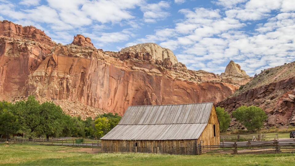 A rustic wooden barn in a grassy field with a backdrop of red rock formations and a clear blue sky.