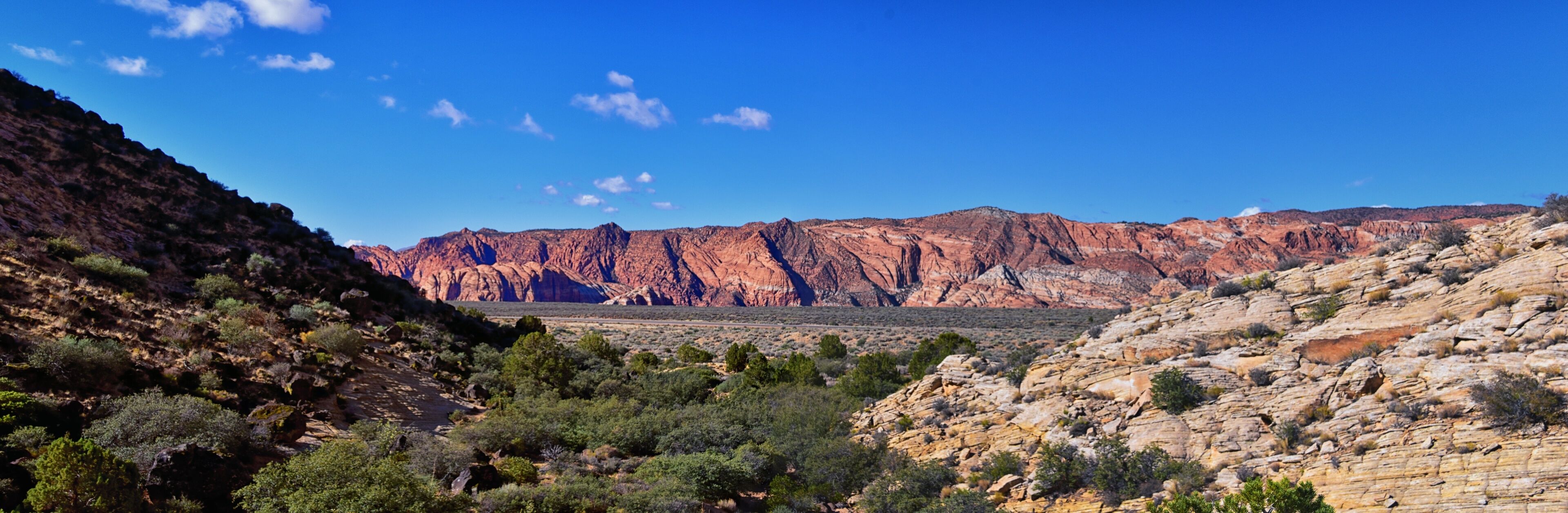 A rugged canyon landscape with layered red rock formations and sparse vegetation.
