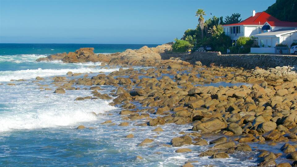 A rocky shoreline with waves crashing against the rocks. In the background, there is a white house with a red roof and a person walking on the beach.