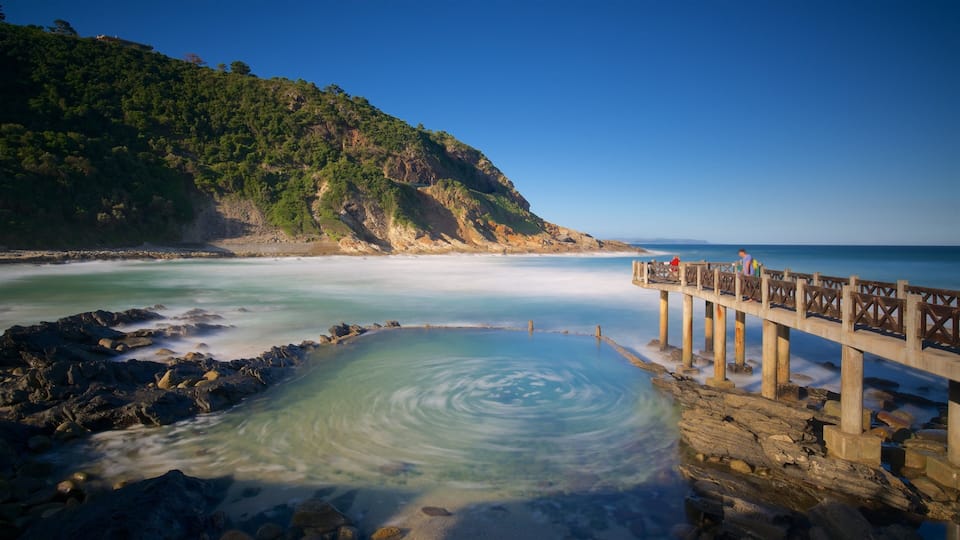 A coastal scene with a wooden pier extending into calm waters, surrounded by rocky shores and a forested hillside.