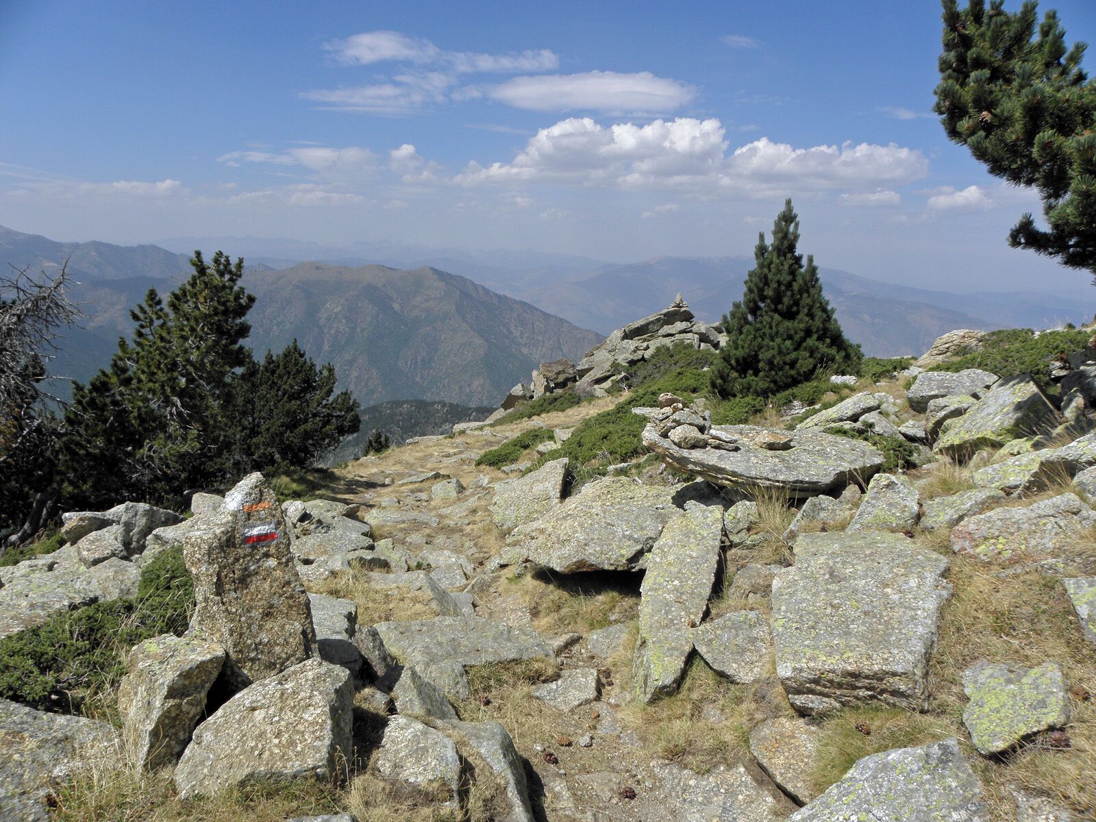 A rocky mountain landscape with sparse vegetation and distant mountains under a clear sky.