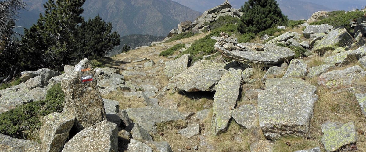 A rocky mountain landscape with sparse vegetation and distant mountains under a clear sky.