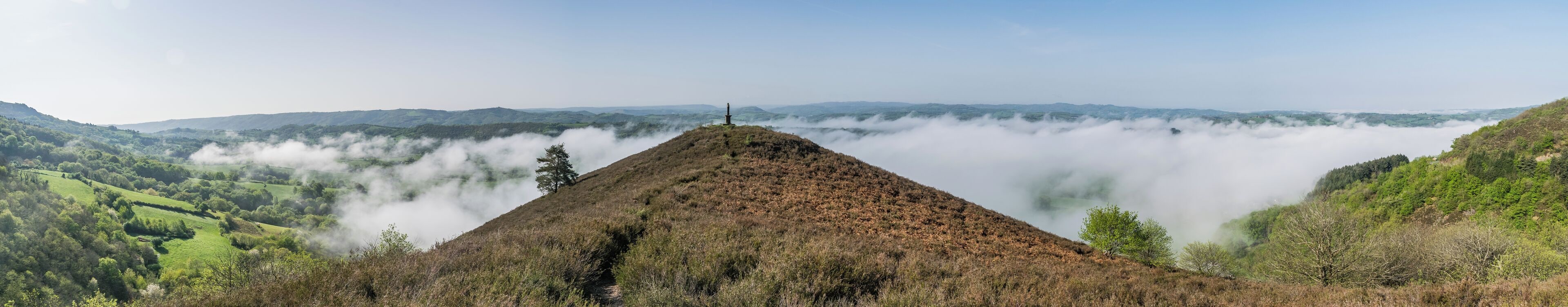 A mountain peak with a monument, surrounded by lush greenery and a valley shrouded in mist.