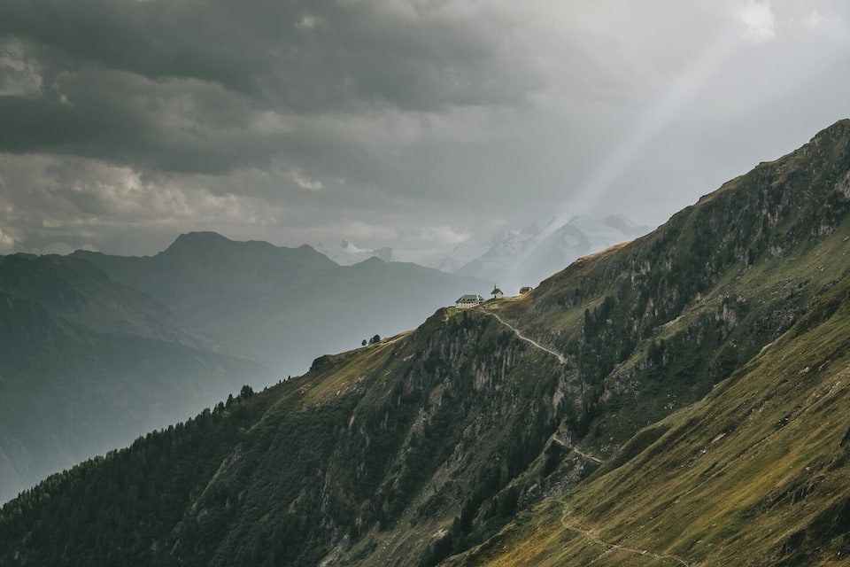 A mountain landscape with a winding path, a small building, and distant peaks under a cloudy sky.