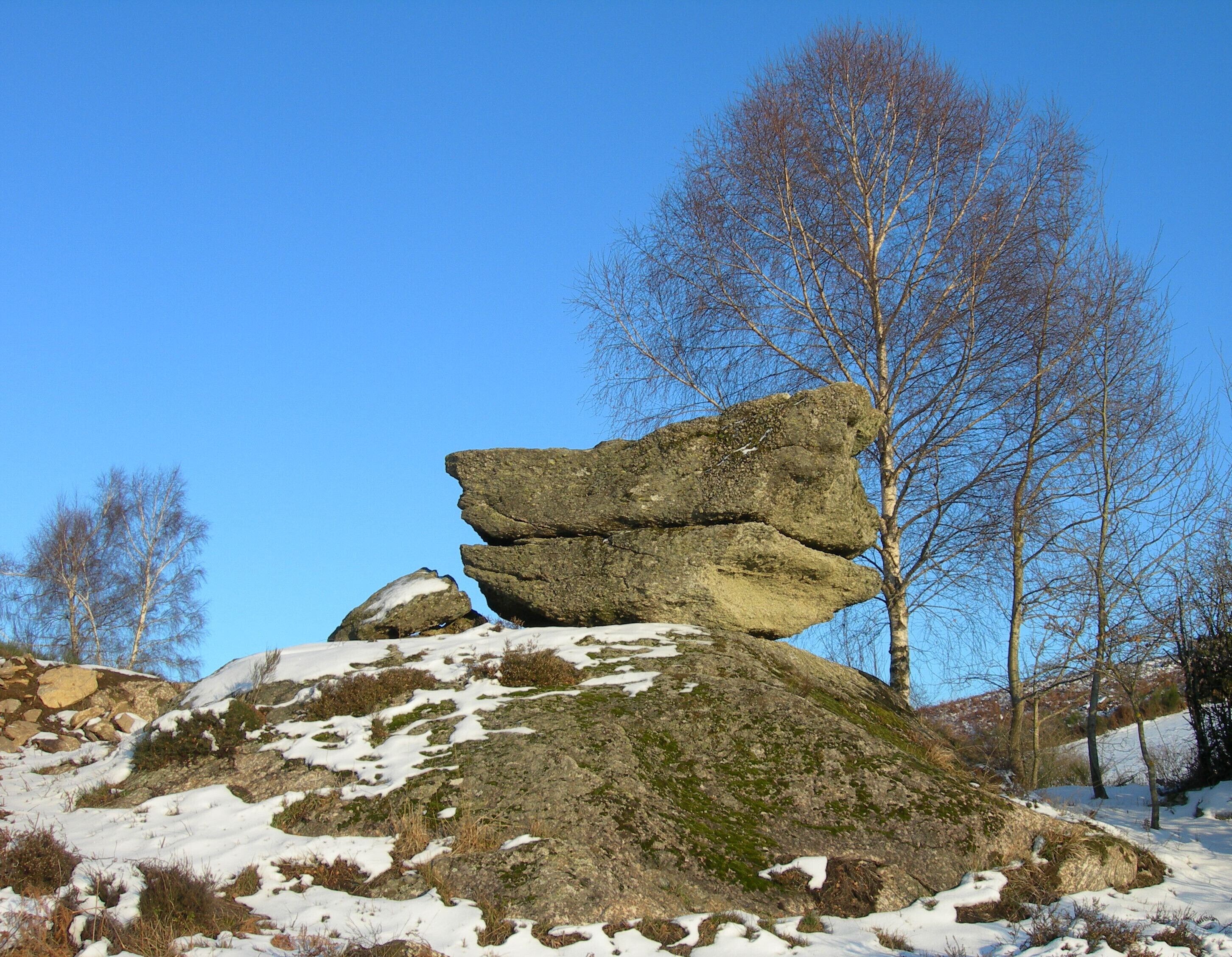 A rock formation with a balanced boulder on top, surrounded by snow and trees.