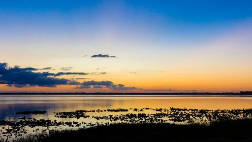 A sunset over a calm lake with reflections of clouds and a distant shoreline.
