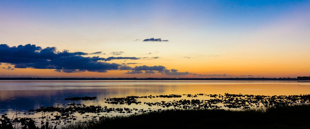 A sunset over a calm lake with reflections of clouds and a distant shoreline.