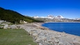 Un rivage rocheux bordé d’un lac aux eaux d’un bleu limpide, une colline boisée et, au loin, des montagnes aux sommets enneigés.