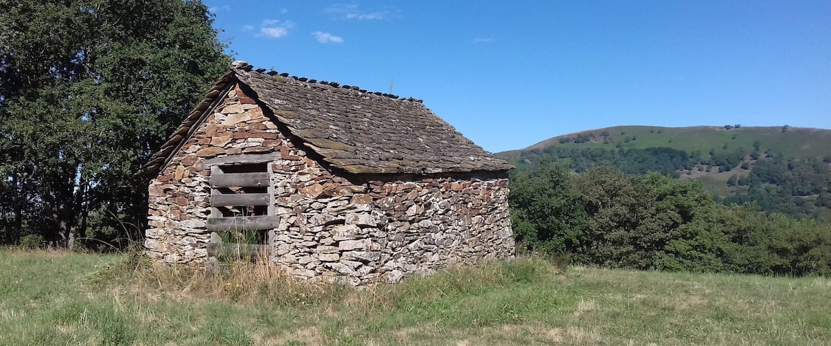 A stone building with a sloped roof in a grassy field, surrounded by trees and hills.